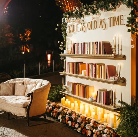 Wedding Ideas: A sofa next to a bookshelf decorated with flowers, candles and text that reads ′Tale as Old as Time′